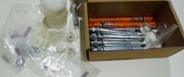 Na stole leżą w pudełku metalowe części do sprzętu medycznego. Obok w woreczkach plastikowe, okrągłe części do sprzętu.