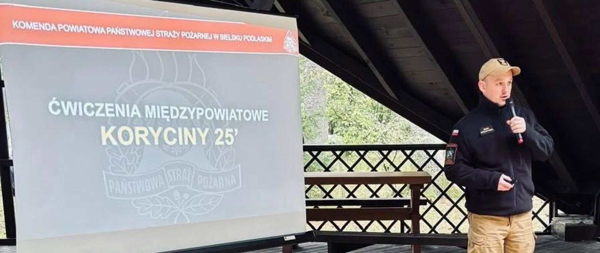 Ćwiczenia pod kryptonimem "KORYCINY 25' - strażak z mikrofonem tłumaczy założenia ćwiczeń , w tle tablica z kryptonimem ćwiczeń