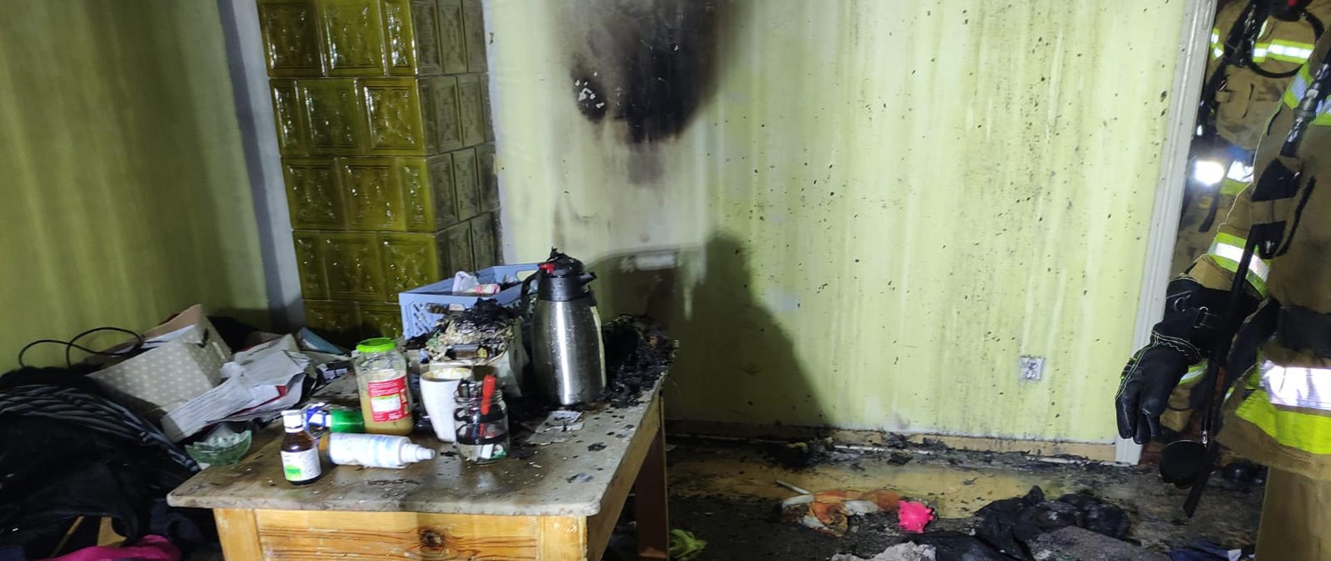 Wnętrze pokoju z osmaloną dymem ścianą oraz resztkami popożarowymi na podłodze.