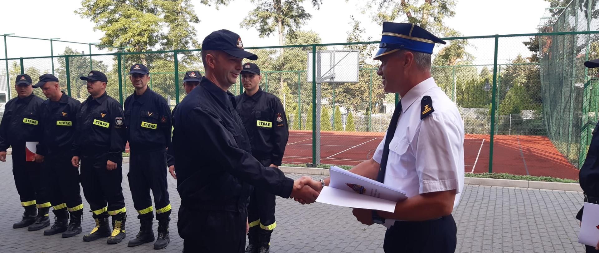 Na pierwszym planie Komendant Powiatowy PSP w Wolsztynie wręcza gratulacje za 20 lat pracy i służby strażakowi wolsztyńskiej Jednostki Ratowniczo-Gaśniczej. Na drugim planie znajdują się ustawieni na zbiórce strażacy.
