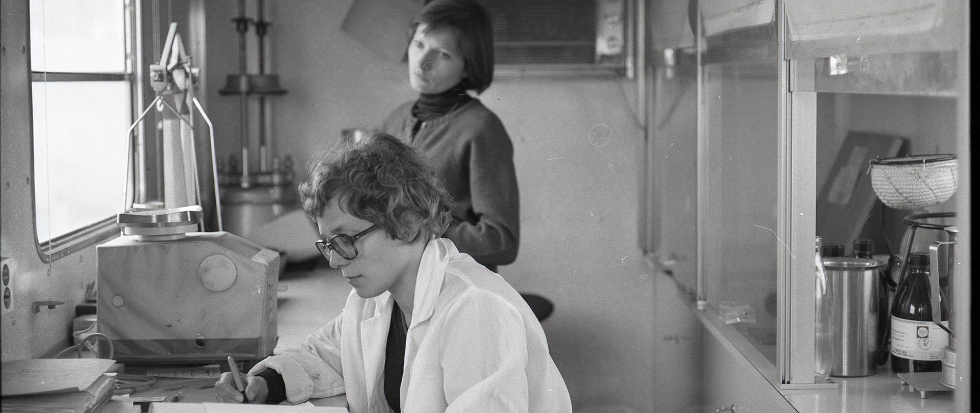 Wąskie pomieszczenie z oknem. Przy ladzie ze sprzętem laboratoryjnym, tyłem do dygestorium, siedzi laborantka w okularach, notuje w zeszycie. Z boku jej pracy przygląda się koleżanka.