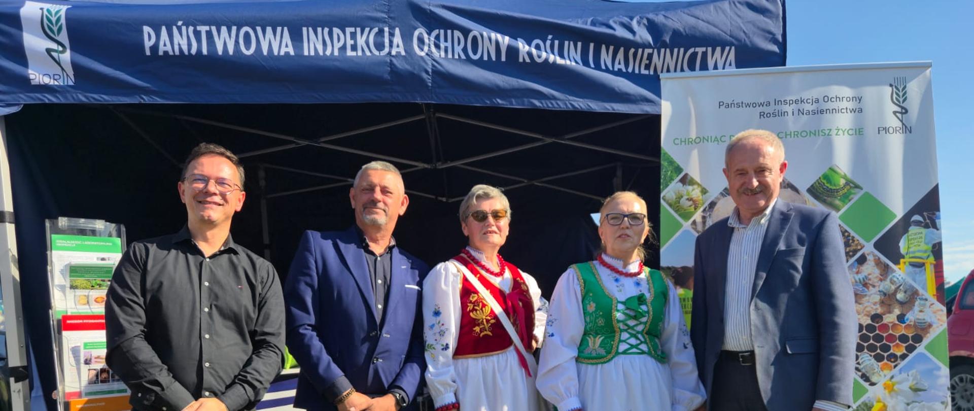 Na zdjęciu widać pięć osób: dwie kobiety w tradycyjnych strojach ludowych, prawdopodobnie z regionu kaszubskiego (na podstawie elementów stroju – hafty, fartuchy, kolorystyka) i trzech mężczyzn ubranych elegancko – w garniturach lub koszulach (przedstawiciele PIORiN) stojących na tle namiotu oraz roll-upu promującego Państwową Inspekcję Ochrony Roślin i Nasiennictwa (PIORiN).