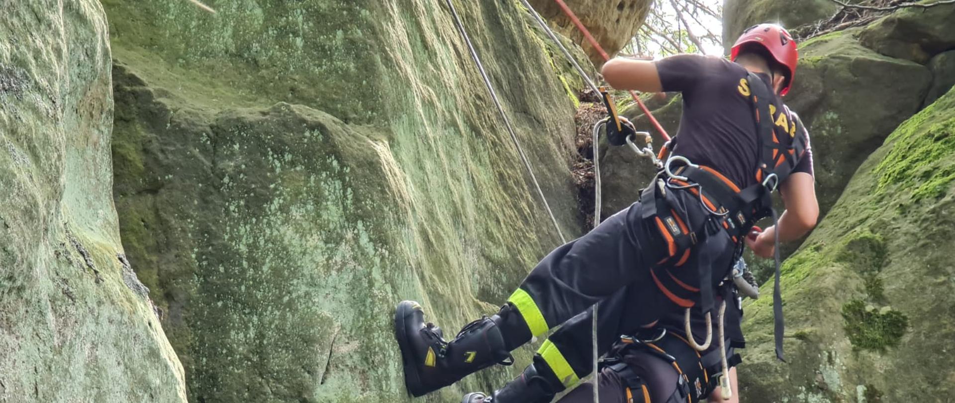 Widoczna skalna formacja po której w asekuracji na linie schodzą ratownicy ubrani z ubrania koszarowe i zabezpieczeni przy pomocy szelek do wspinaczki. 