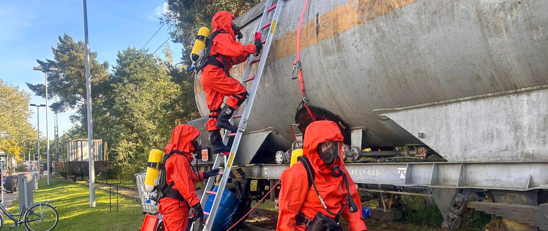 Trzech ratowników w pomarańczowych kombinezonach ochronnych przy cysternie kolejowej. Jeden z nich schodzi po drabinie, dwaj pozostali pracują przy zbiorniku. Widoczne są węże, butle oddechowe i sprzęt zabezpieczający.