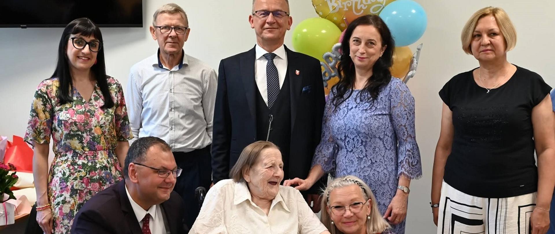 W centrum, przy stole , na którym stoi duży tort urodzinowy ze świeczkami w kształcie liczby100, siedzi starsza kobieta. Po jej lewej stronie przykucnął mężczyzna w garniturze, a po prawej – kobieta. Za nimi stoją trzy kobiety i dwóch mężczyzn. W tle znajdują się kolorowe balony i dekoracja z napisem „Happy Birthday”.