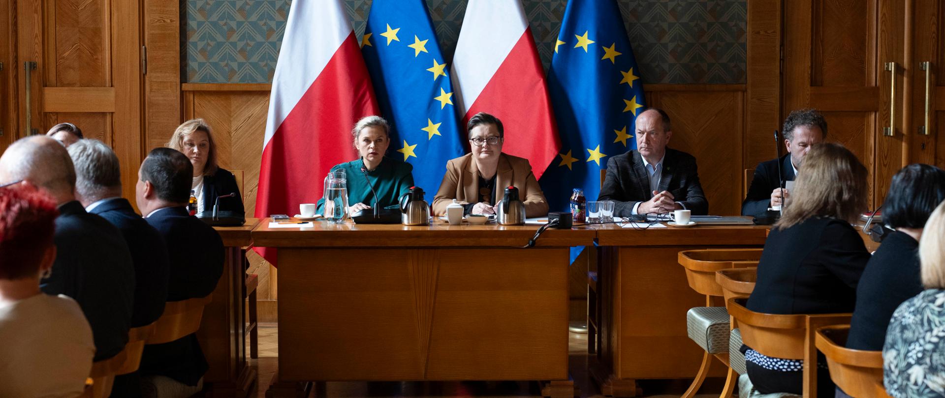 Szerokie ujęcie sali konferencyjnej, gdzie grupa osób siedzi przy dwóch rzędach stołów. W tle, za centralnym stołem, siedzą cztery osoby na tle polskiej i unijnej flagi.