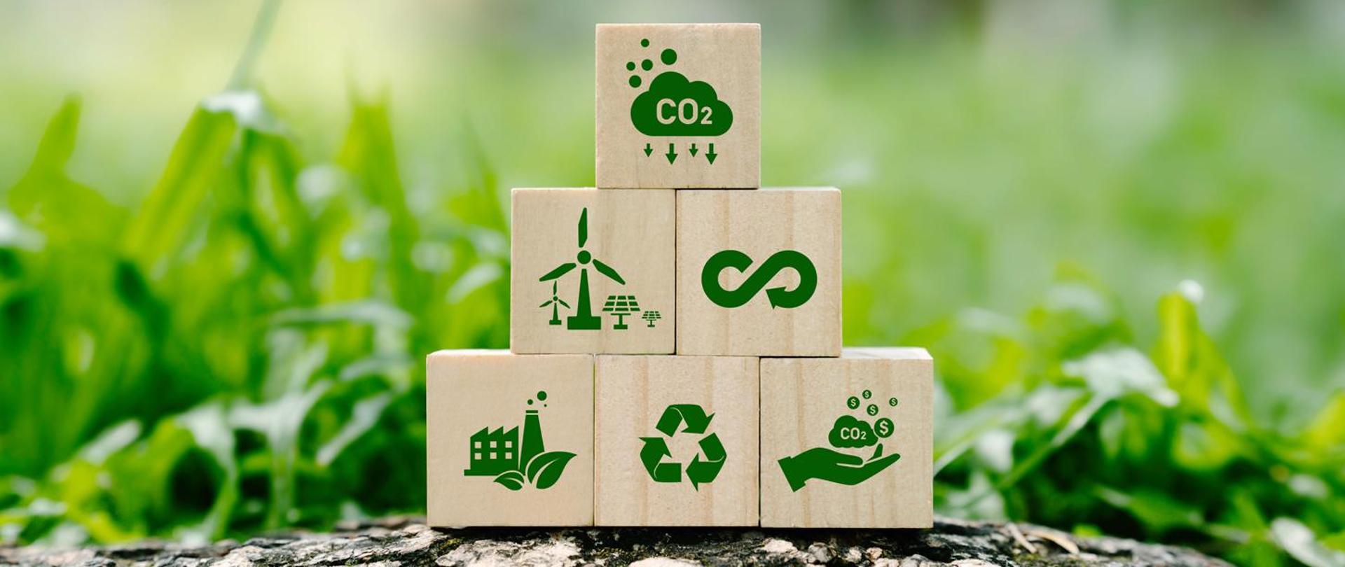 Na środku ułożone w piramidę drewniane klocki z zielonym ikonkami. W dolnym pasku od lewej ikonka elektrowni, druga ikonka 3 strzałki ułożone w trójkąt trzecia ikonka dłoń a nad nią chmurki z napisem CO2i ikonami dolara. W drugim rzędzie piramidy ikonka wiatraków wiatrowych oraz fotowoltaiki, kolejna ikonka znak nieskończoności. Na górze piramidy ikonka chmurki z napisem CO2 pod chmura strzałki skierowane w dół. W tle piramidy widać niewyraźnie trawę.