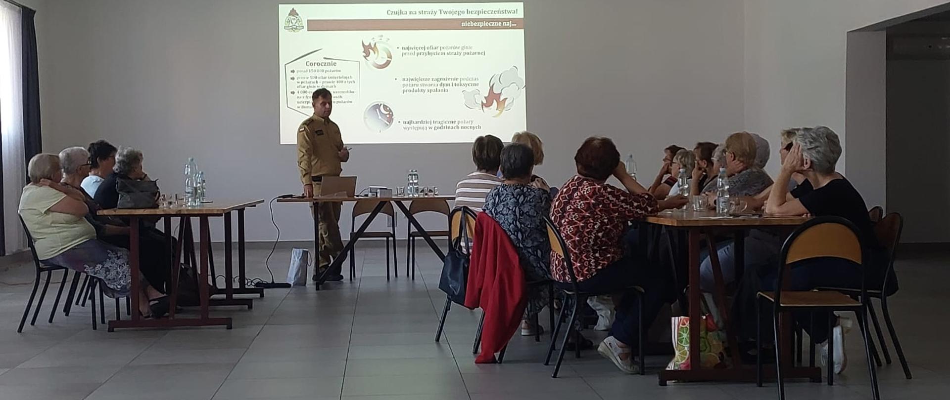 Seniorzy siedzący w ławkach i słuchający centralnie ustawionego funkcjonariusza PSP z Węgrowa przedstawiającego prezentacje o czujkach.
