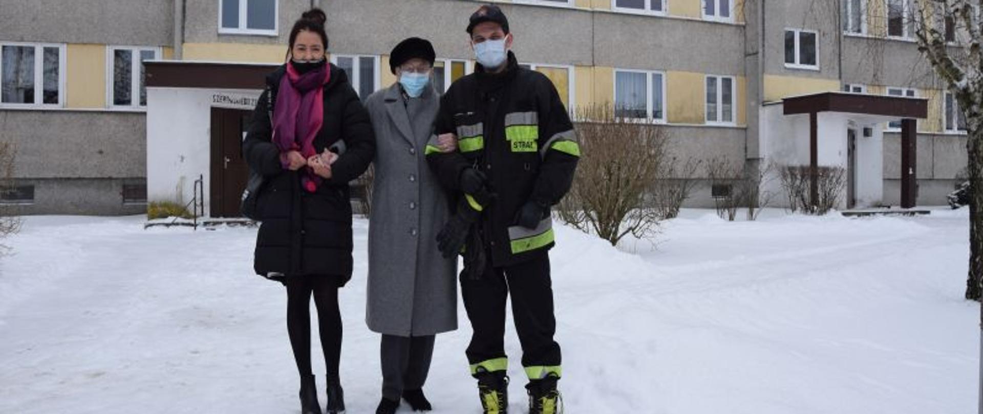 Na zdjęciu widać strażaka , którego trzyma pod rękę starsza kobieta, która udaje się przy pomocy strażaka i drugiej kobiety do samochodu.