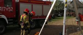 Dwóch strażaków Państwowej Straży Pożarnej przygotowań do wejścia do studni w celu wydobycia źrebaka, który do niej wpadł. Strażacy wyposażeni w szelki alpinistyczne. Obok rozstawiony jest trójnóg ewakuacyjny. W tle samochód ratowniczo-gaśniczy.