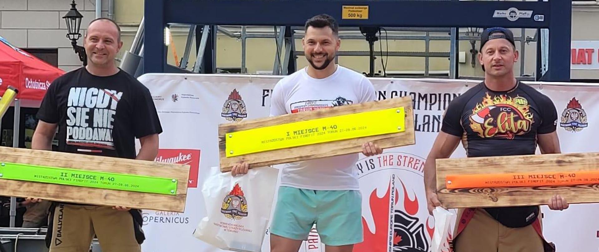 Zdjęcie przedstawia zawodników stojących na podium po zawodach FIREFIT 2024 w Toruniu, trzymają w rękach nagrody w tle znajduje się wieża i budynki mieszkalne.