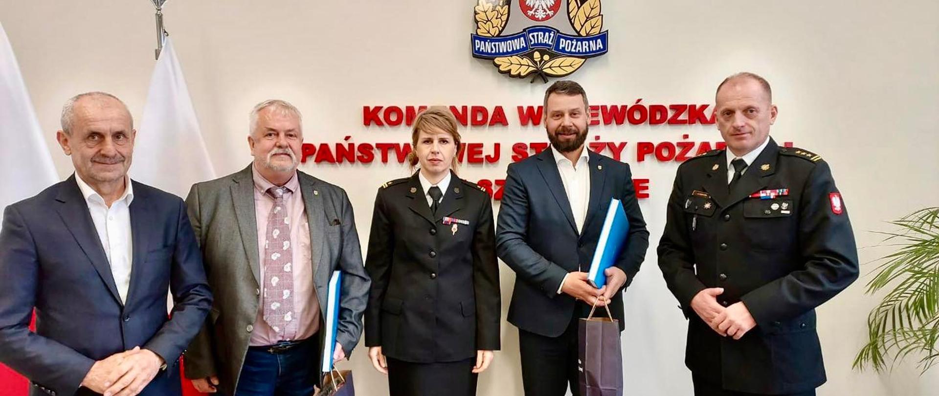 Na zdjęciu widać 5 osób stojących na tle ściany pamiątkowej KW PSP Szczecin. Wśród osób znajdują się goście z Niemiec,
Zachodniopomorski Komendant Wojewódzki PSP st. bryg. Jarosław Tomczyk, druh Marek Kowalski przedstawiciel ZOSP RP oraz specjalista ds. współpracy transgranicznej
bryg. Agnieszka Beśka-Mazurek.
