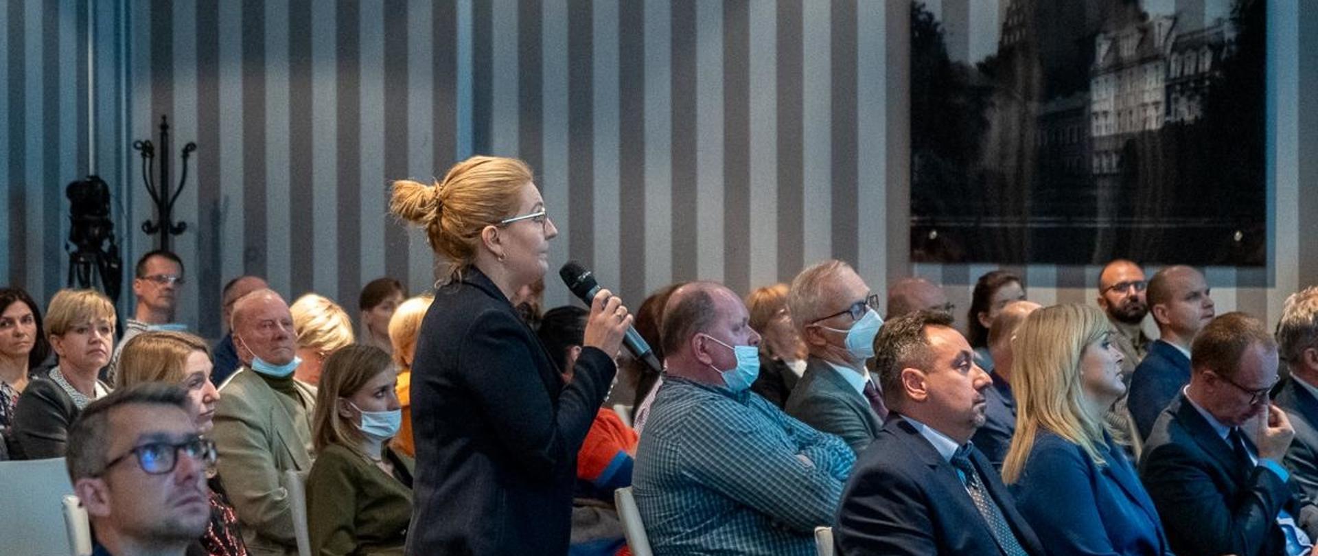 Kobieta w koku i okularach, trzyma mikrofon i stoi pośrodku widowni. Wszyscy obserwują prezentację