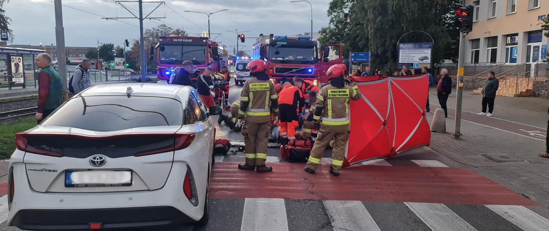 Ratownicy medyczni i strażacy udzielają pomocy osobom poszkodowanym na przejściu dla pieszych. W tle widoczne pojazdy straży pożarnej i karetki pogotowia. Po lewej stronie pojazd osobowy.