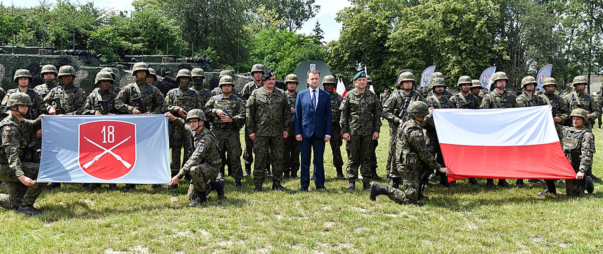 Minister Mariusz Błaszczak stoi z żołnierzami Wojska Polskiego podczas wizyty w 18. Dywizji Zmechanizowanej. Po prawej stronie żołnierze trzymają flagę z oznaką Dywizji. Po prawej flagę Polski. W tle sprzęt wojskowy i roślinność.