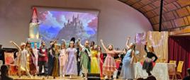 Grupa dzieci w bajkowych strojach (księżniczki, rycerze, wróżki) stoi na scenie w dynamicznych pozach z uniesionymi rękami. W tle duża projekcja zamku na wzgórzu. Widoczni widzowie na widowni.