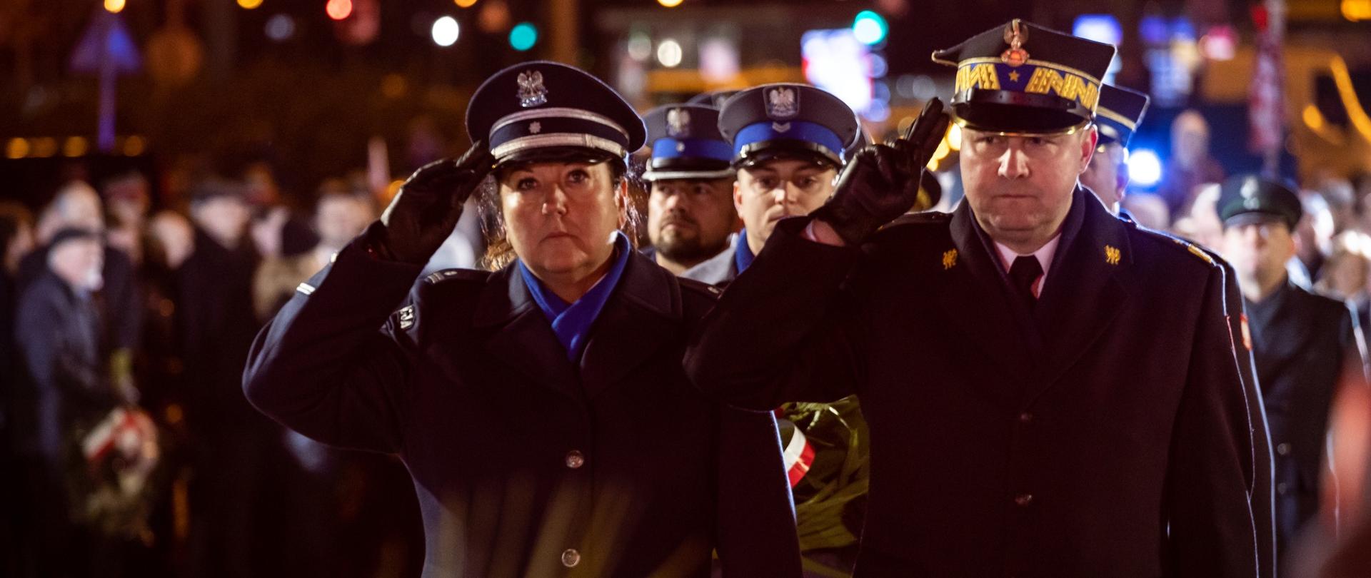 strażak w płaszczu i rogatywce salutuje, obok stoi kobieta policjant stoi w płaszczu i rogatywce, 