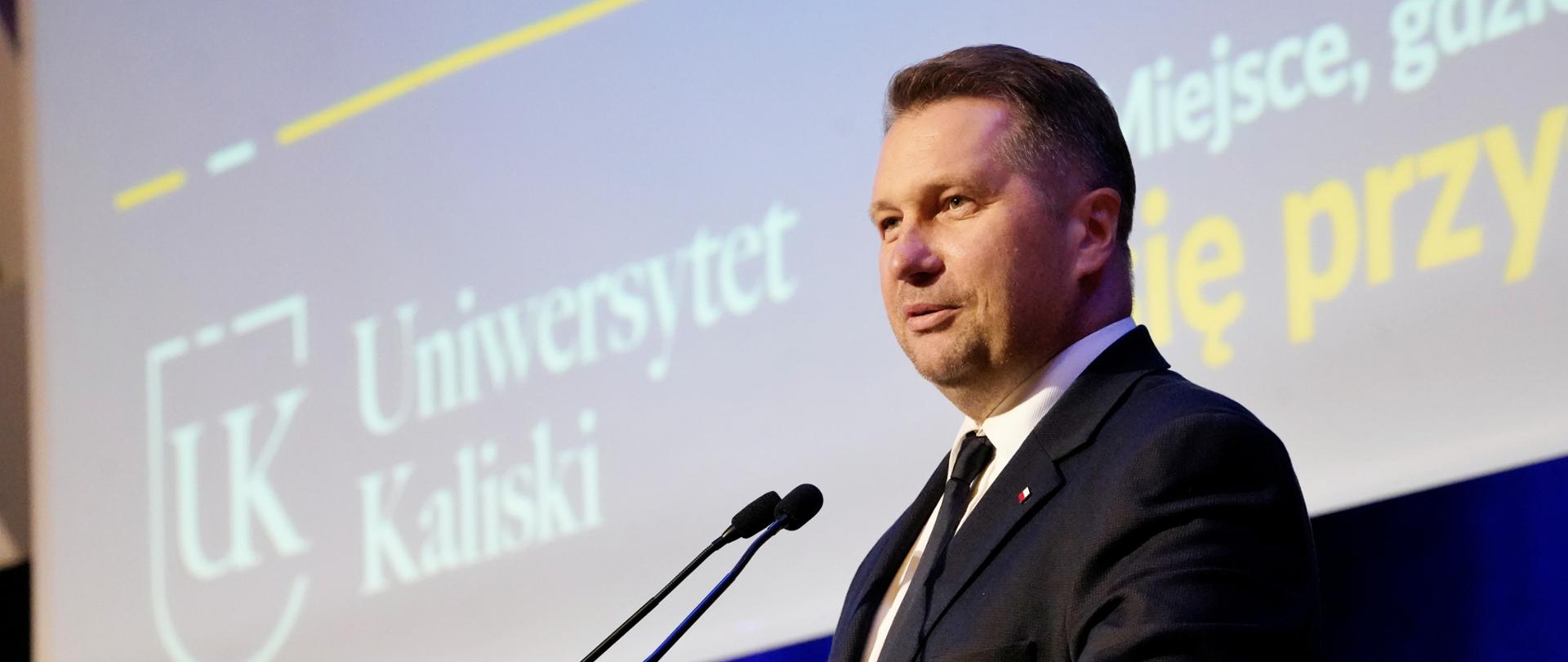 Minister Czarnek stoi za drewniana mównicą i mówi do dwóch małych mikrofonów.