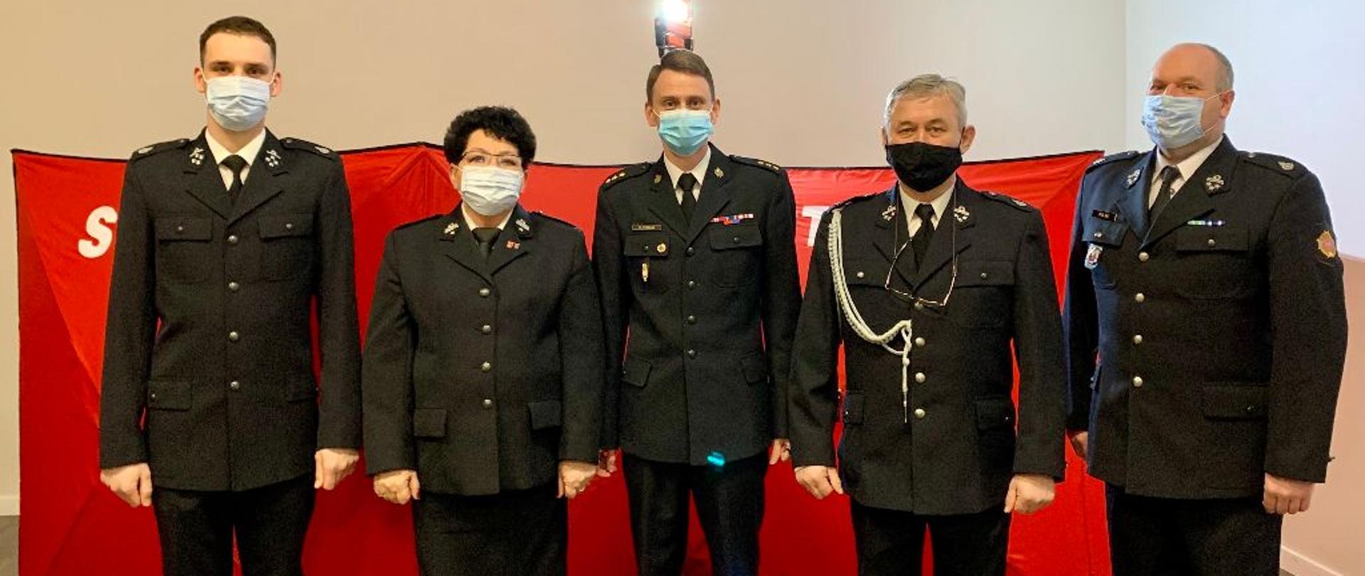 Zdjęcie przedstawia pięć osób stojących na tle parawanu do zabezpieczenia akcji ratowniczych. Od lewej stoją: Prezes OSP Wielkie Radowiska, Prezes Zarządu Gminnego Związku OSP Dębowa Łąka, Zastępca Komendanta Powiatowego PSP w Wąbrzeźnie, Komendant Gminny OSP Dębowa Łąka, Naczelnik OSP Wielkie Radowiska. Osoby są ubrane w mundury wyjściowe, na twarzach mają założone maseczki ochronne.