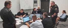 Sztab Opolskiego Komendanta Wojewódzkiego PSP w Opolu podczas ćwiczeń na zakładzie dużego ryzyka wystąpienia poważnej awarii przemysłowej. Funkcjonariusze w ubraniach służbowych przy stole wewnątrz sali sztabowej wydziału operacyjnego KW PSP w Opolu. W tle tablica suchościeralna z zanotowanym schematem łączności.