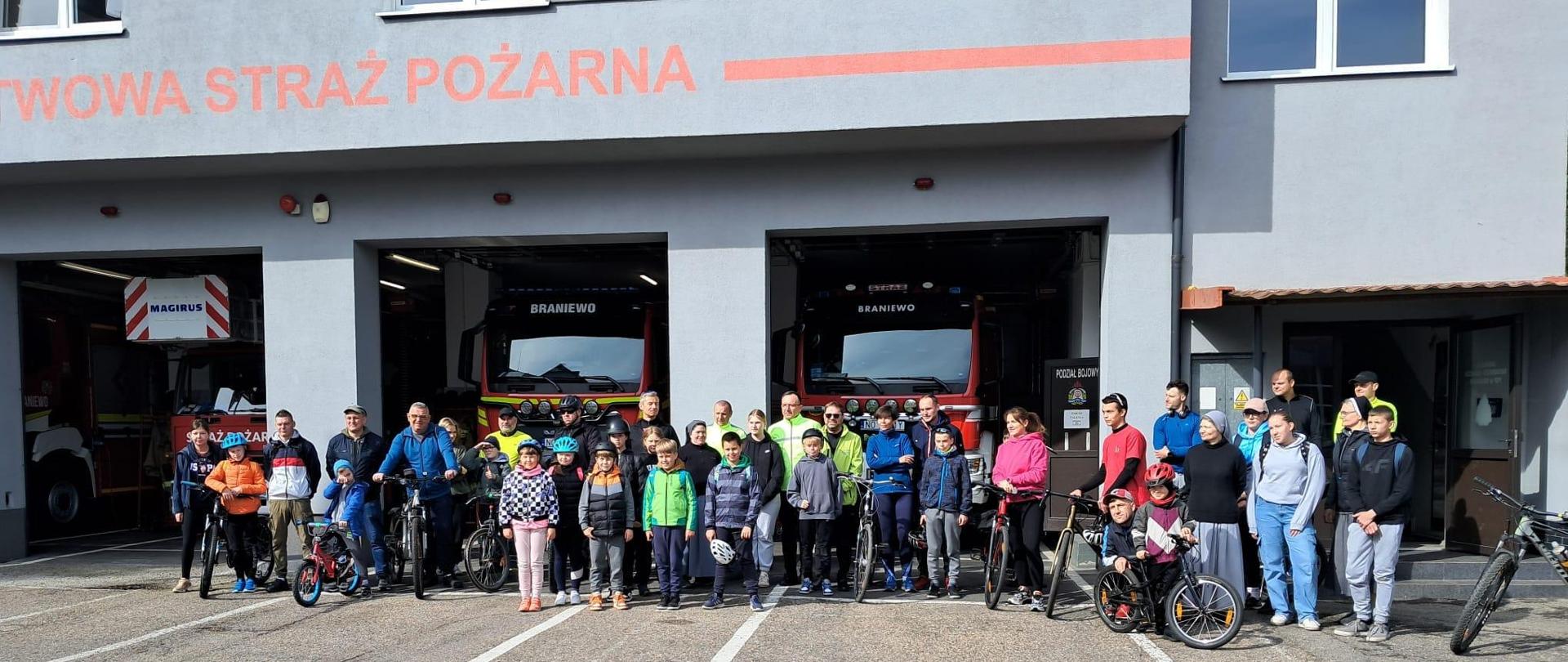Na tle remizy PSP stoi duża grupa osób, uczestników rajdu rowerowego.