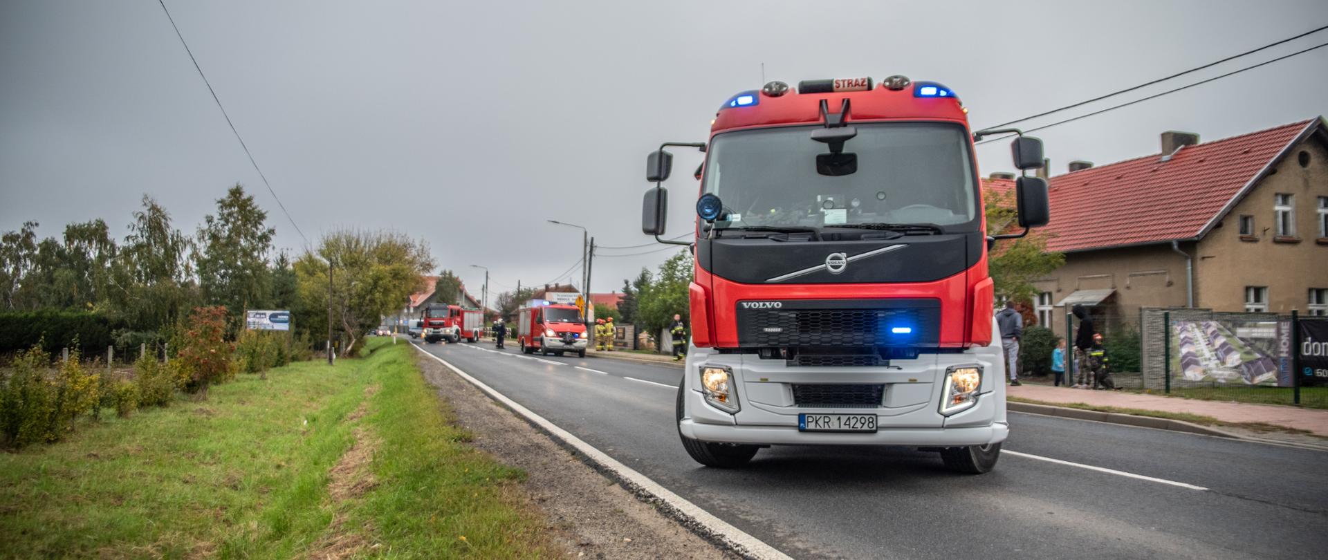 Pojazdy straży pożarnej blokują drogę, w tle strażacy realizujący działania ratownicze. W pobliżu miejsca zdarzenia duża ilość osób postronnych. Pojazdy mają włączone sygnały błyskowe. 