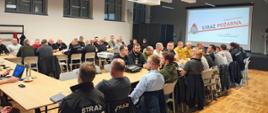 Duże spotkanie w sali konferencyjnej: kilkudziesięciu uczestników, w tym strażacy i osoby cywilne, siedzi przy długich stołach i prowadzi rozmowy. Na końcu sali widoczny jest ekran z prezentacją „Państwowa Straż Pożarna”. Wnętrze jest nowoczesne, z wysokim sufitem, dużymi oknami i zawieszonym oświetleniem.
