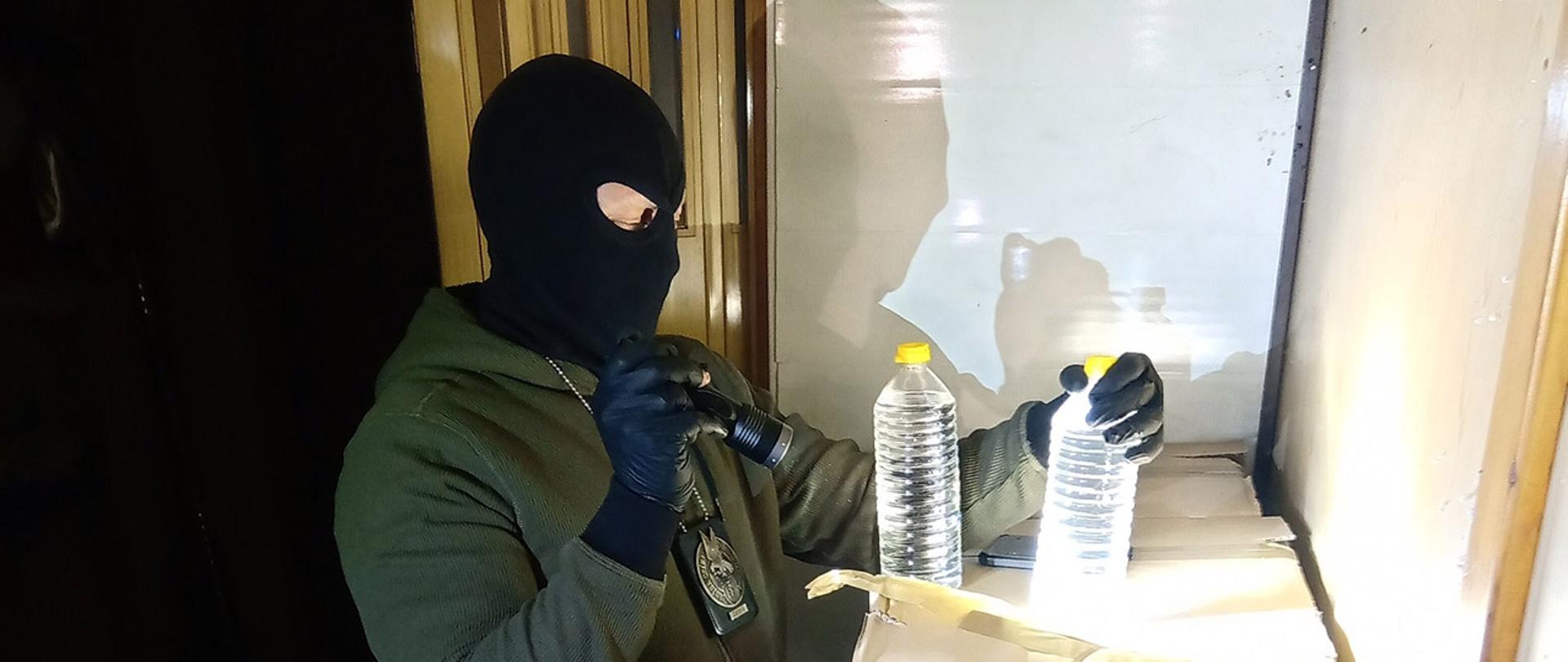 Funkcjonariusz KAS w kominiarce i z latarką podczas inspekcji butelek z nielegalnym alkoholem.