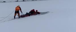 Strażacy ubrani w suche skafandry. Jeden z nich znajduje się na saniach lodowych, natomiast drugi przytrzymuje sanie które znajdują się na zamarzniętym zalewie. Strażak leżący na saniach próbuje wyciągnąć osobę znajdującą się w wodzie.