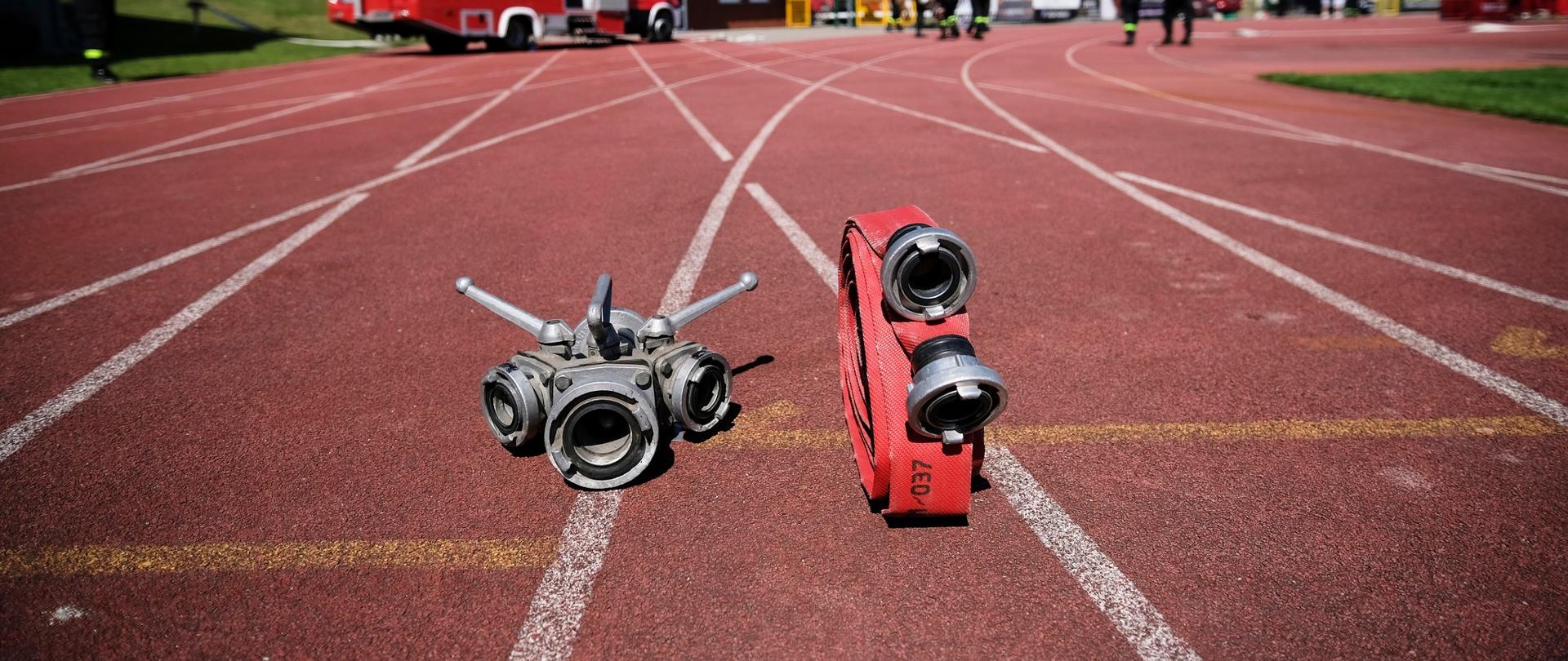 Tartanowa bieżnia stadionu w kolorze czerwonym, na tartanie ustawione rozdzielacz i wąż pożarnicy. Ostrość zdjęcia skupiona na tych przedmiotach.