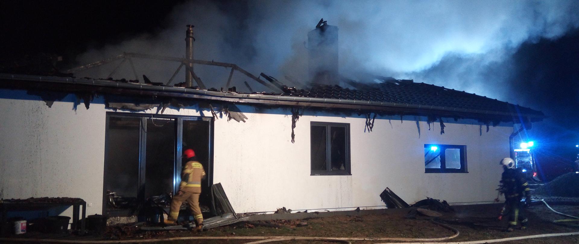 Zdjęcie przedstawia dom ze spalonym dachem w porze nocnej. Przed domem jest 2 strażaków i rozwinięte linie gaśnicze.
