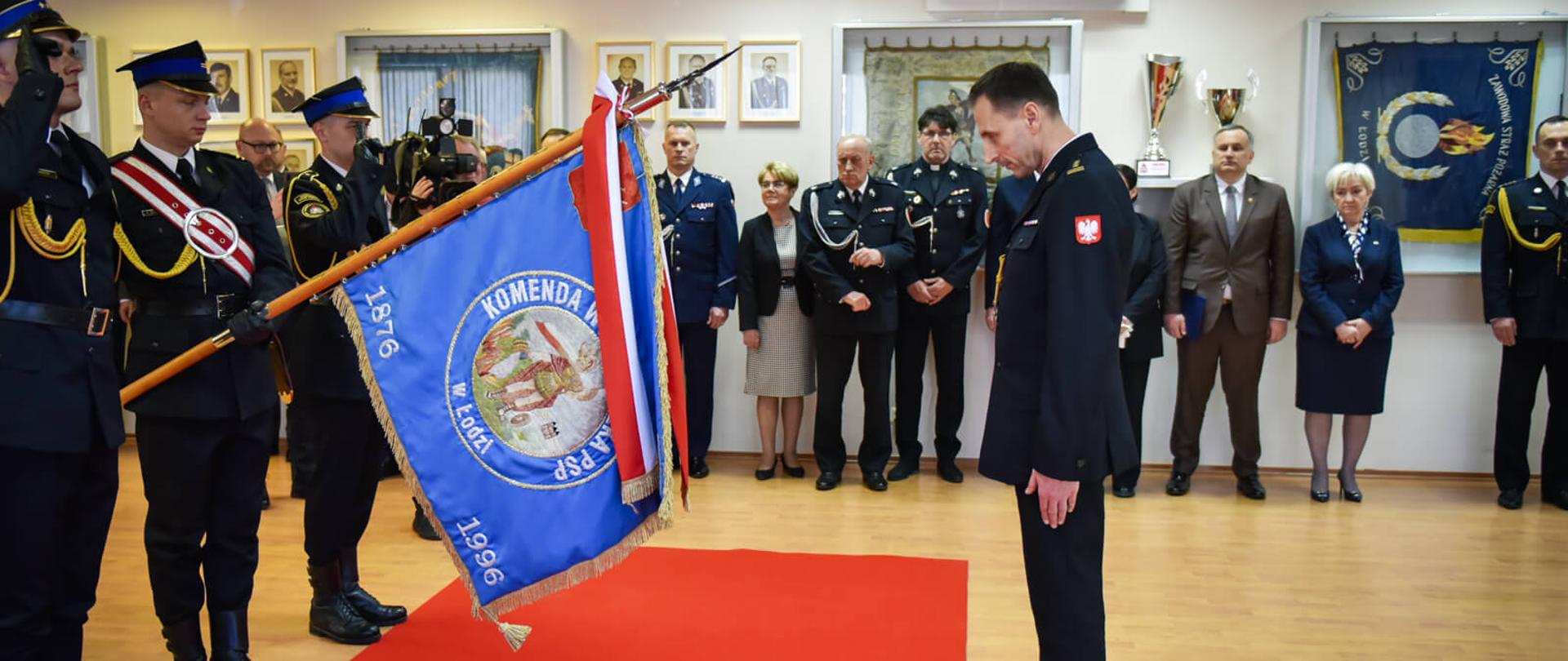 Nowy komendant wojewódzki PSP pochyla głowę przed niebieskim sztandarem Komendy Wojewódzkiej PSP w Łodzi, za nim zgromadzeni goście i funkcjonariusze.