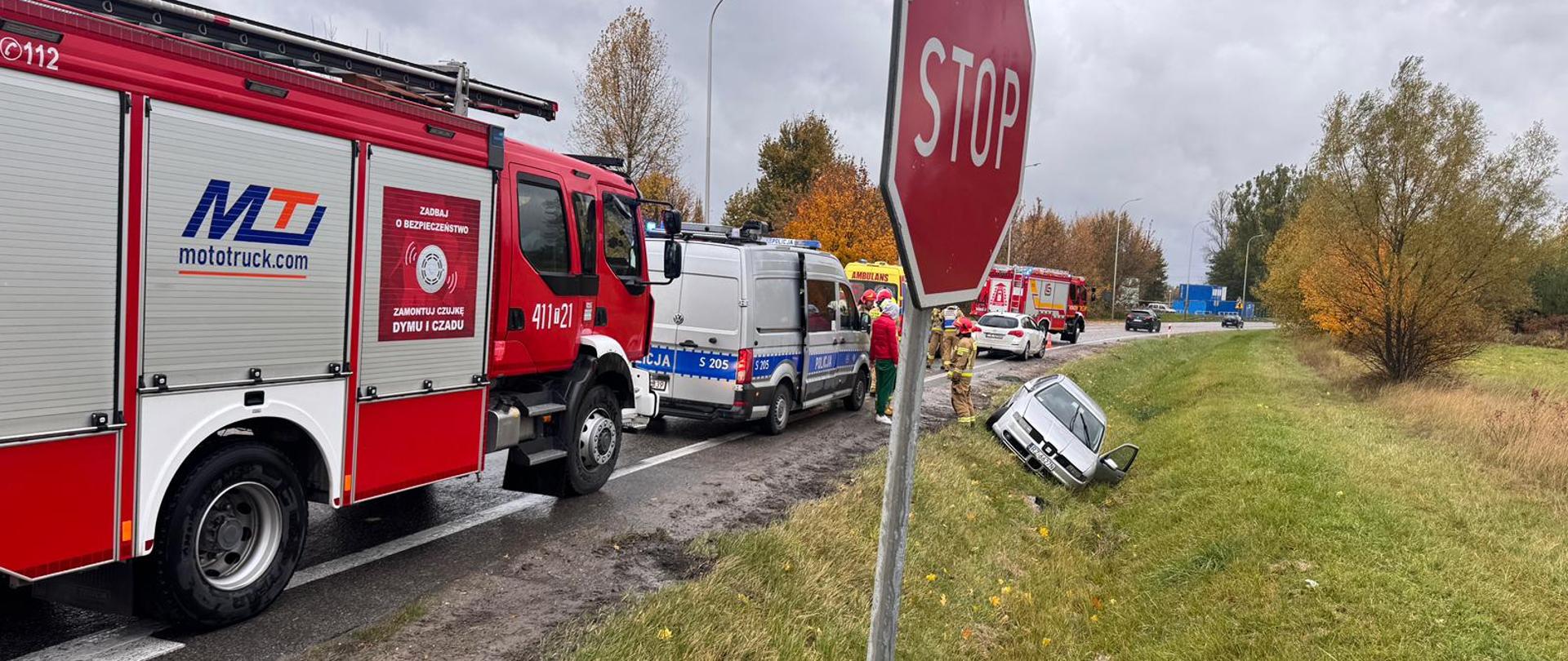 Zdjęcie przedstawia na pierwszym planie wóz strażacki oraz radiowóz policyjny. W tle uszkodzony pojazd osobowy po wcześniejszym zdarzeniu w przydrożnym rowie. Za nimi samochód ratowniczo-gaśniczy, karetka pogotowia w kolorze żółtym.
