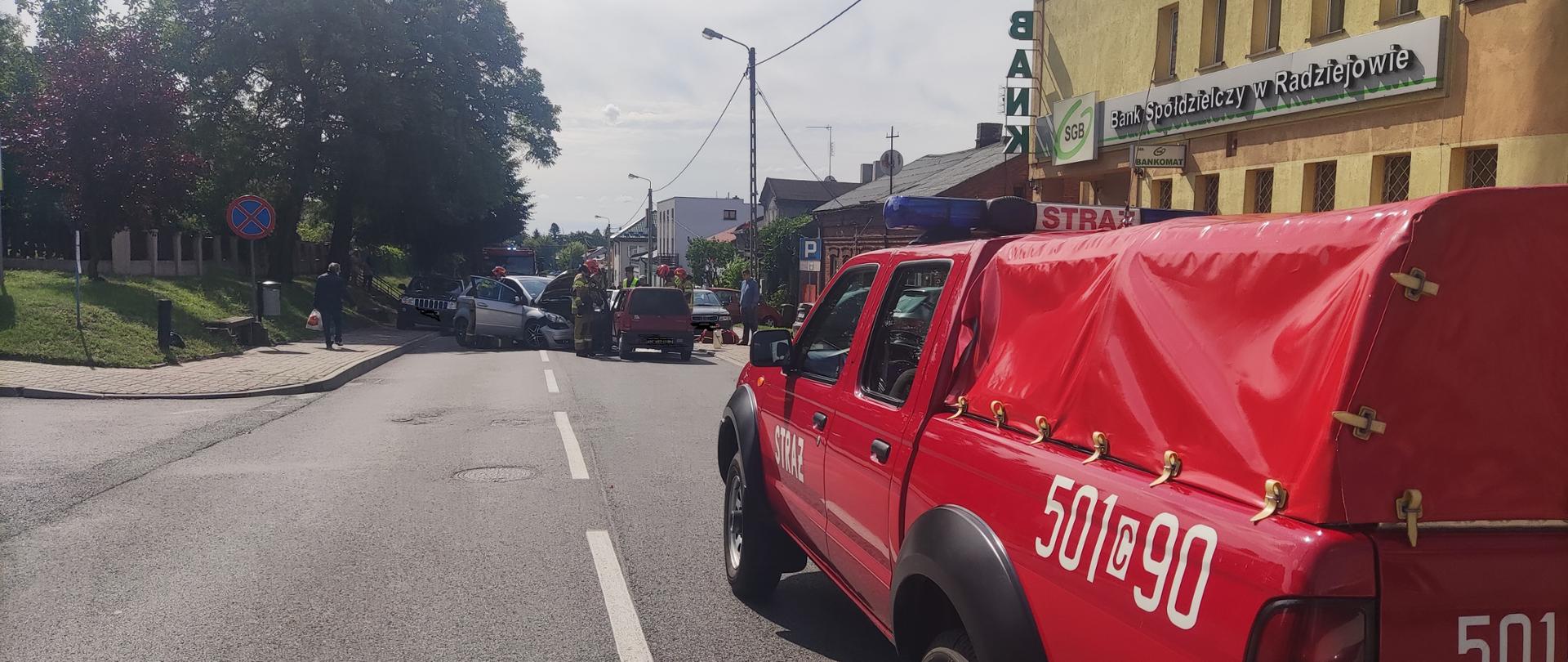 Zdjęcie przedstawia pas drogowy ulicy Kościuszki, na której znajdują się pojazdy służb ratowniczych (dwa wozy straży pożarnej) strażaków i policjantów podczas czynności służbowych po kolizji drogowej dwóch pojazdów osobowych.