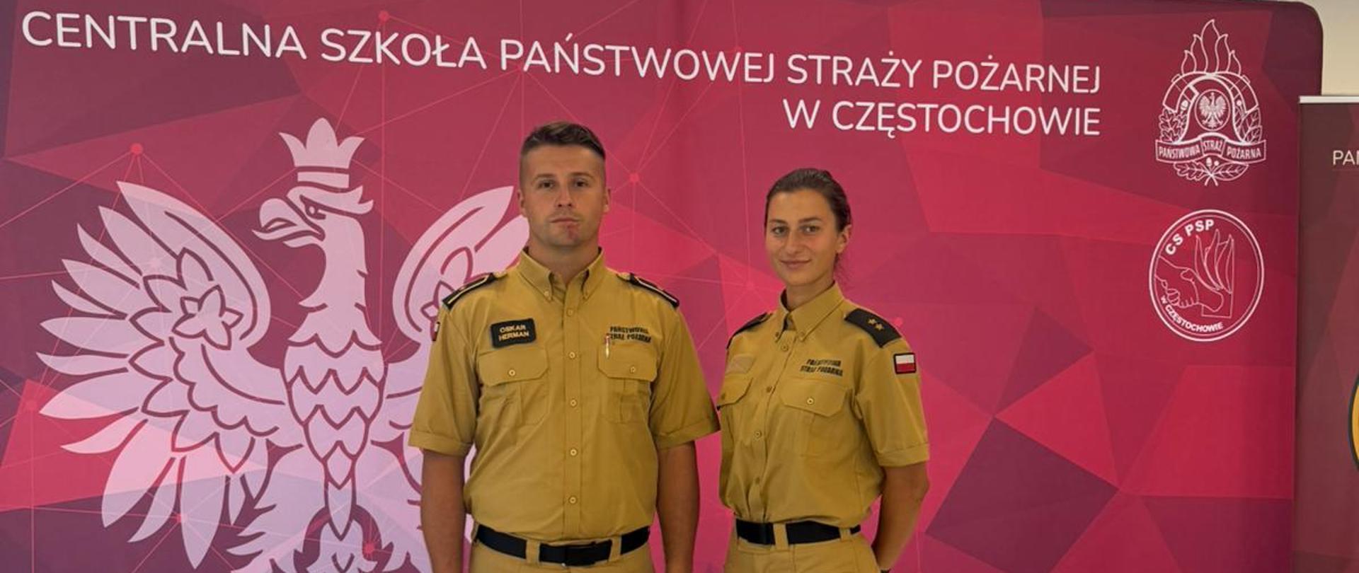 Od lewej stojący w mundurze koloru piaskowego aspirant CS PSP obok niego stoi w mundurze koloru piaskowego Pani młodszy kapitan CS PSP, za nimi czerwony baner z umieszczonym na górze napisem Centralna Szkoła Państwowej Straży Pożarnej, poniżej napisu po lewej wielkie godło po prawej od góry logo PSP oraz CS PSP