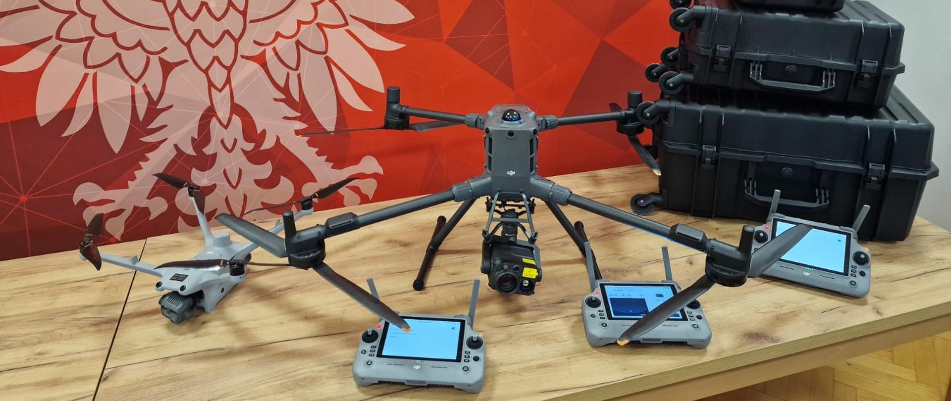 Drony ratownicze z kamerami i kontrolerami, umieszczone na stole przed ścianką Komendy Powiatowej Państwowej Straży Pożarnej w Kępnie; obok widoczne są czarne walizki transportowe na sprzęt.