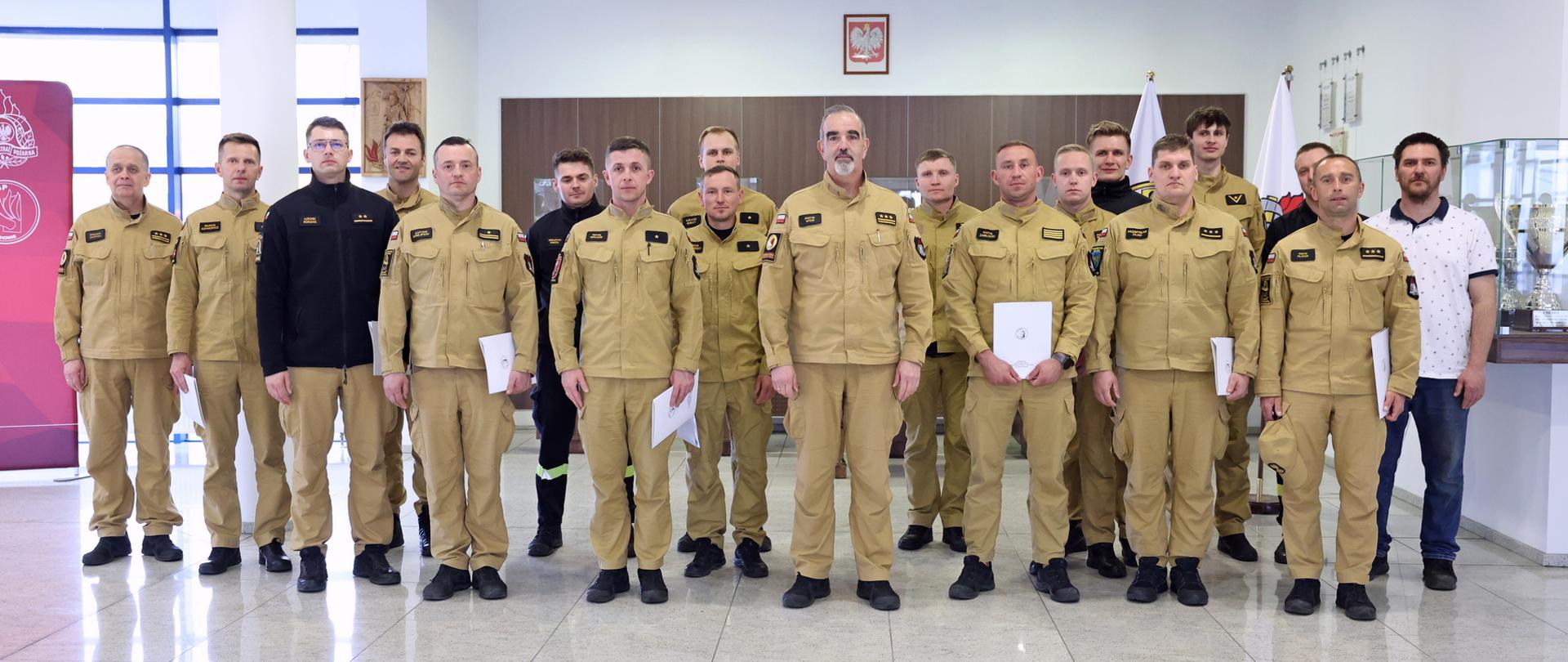 Pamiątkowe zdjęcie grupy funkcjonariuszy PSP kończących szkolenie (16 osób) w towarzystwie Zastępcy Komendanta Centralnej Szkoły PSP, oficera CS PSP oraz mężczyzny będącego jednym z wykładowców podczas szkolenia