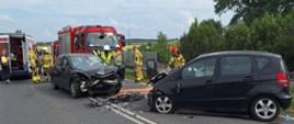 Zdjęcie przedstawia miejsce czołowego zderzenia dwóch samochodów osobowych na środku drogi dwukierunkowej. Czarny samochód po prawej stronie ma poważnie uszkodzony przód i wbity przód drugiego, ciemniejszego auta, również z mocno zniszczonym przodem. Na jezdni widoczne są rozrzucone odłamki, a wokół pojazdów stoją strażacy w żółtych mundurach oraz ratownicy medyczni. Po lewej stronie zdjęcia znajdują się czerwony wóz strażacki i ambulans z otwartymi drzwiami. W tle widać pas zieleni z drzewami oraz niebo z lekkim zachmurzeniem.