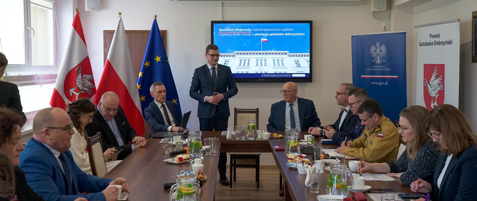 Grupa osób siedzących przy stole. W części prezydialnej stołu stoi wojewoda. Za nim ekran telewizyjny i flagi. 