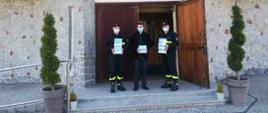 Zdjęcie przedstawia trzech strażaków ochotniczej straży pożarnej w umundurowaniu koszarowym stojących przy wejściu do kościoła. Strażacy trzymają w ręku po kilka opakowań z maseczkami. Wszyscy posiadają na twarzy maseczkę.