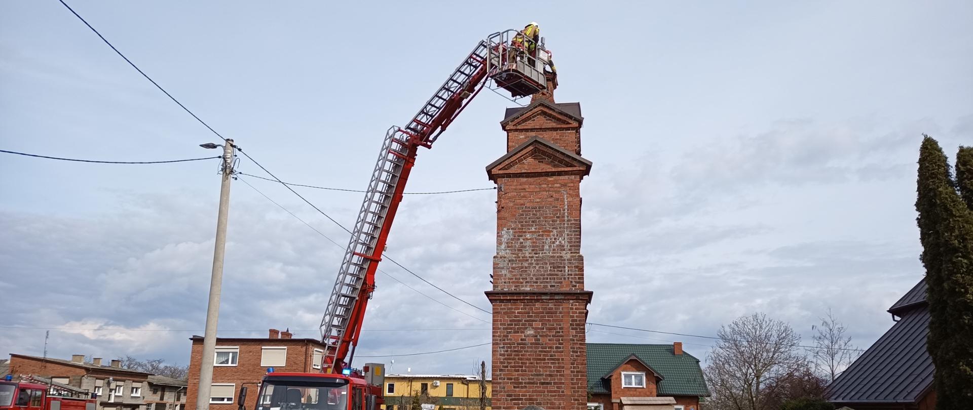 Na zdjęciu rozłożony podnośniki. Podnośnik na samochodzie Volvo, czerwony, sygnalizacja ostrzegawcza. Obok budowla z czerwonej cegły. Zachmurzenie lekko. Po lewej ratownik w ubraniu specjalnym. Plac asfaltowy. W tle budynki mieszkalne.