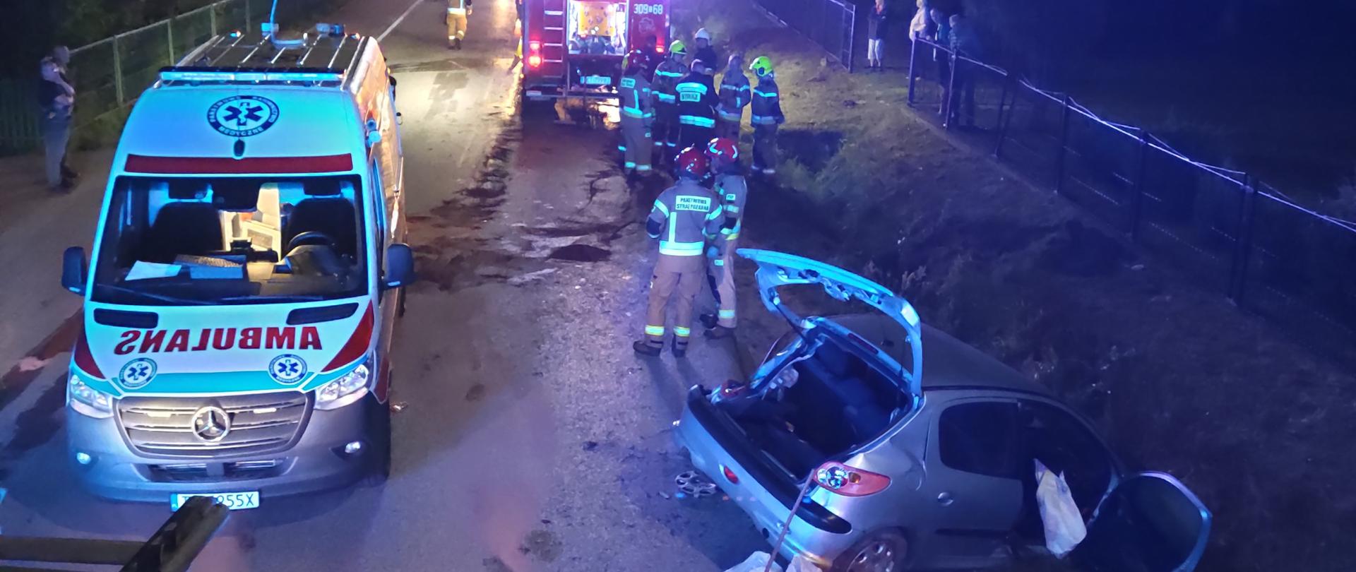 Zdjęcie przedstawia miejsce wypadku: z lewej strony ambulans, a z prawej, częściowo w rowie zniszczony samochód. W głębi samochód pożarniczy i grupa ratowników. 