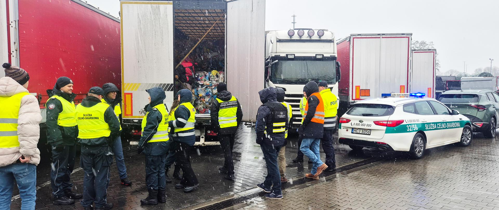 Grupa funkcjonariuszy w żółtych kamizelkach odblaskowych kontroluje otwartą ciężarówkę w deszczowy dzień; wewnątrz widać liczne zapakowane worki, a obok stoi radiowóz Służby Celno-Skarbowej z włączonymi sygnałami. Blisko inne ciężarówki.