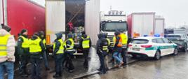 Grupa funkcjonariuszy w żółtych kamizelkach odblaskowych kontroluje otwartą ciężarówkę w deszczowy dzień; wewnątrz widać liczne zapakowane worki, a obok stoi radiowóz Służby Celno-Skarbowej z włączonymi sygnałami. Blisko inne ciężarówki.