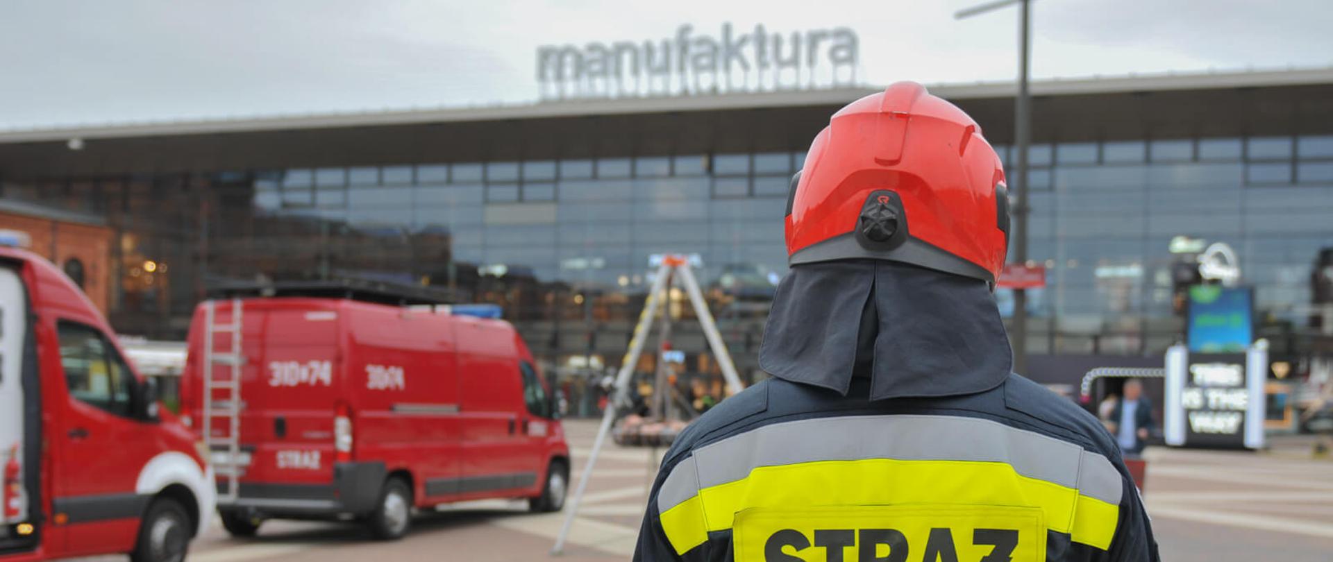 na zdjęciu strażak tyłem do fotografującego po lewej dwa samochody strażackie a w tle budynek z napisem manufaktura