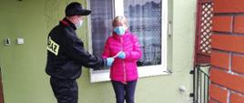 Zdjęcie przedstawia strażaka OSP, który wręcza kobiecie ulotkę dotyczącą szczepień przeciwko COVID-19. Zdjęcie wykonane jest przed wejściem do budynku mieszkalnego. Na ścianie budynku znajduje się numer domu: 4 oraz napis: sołtys. 