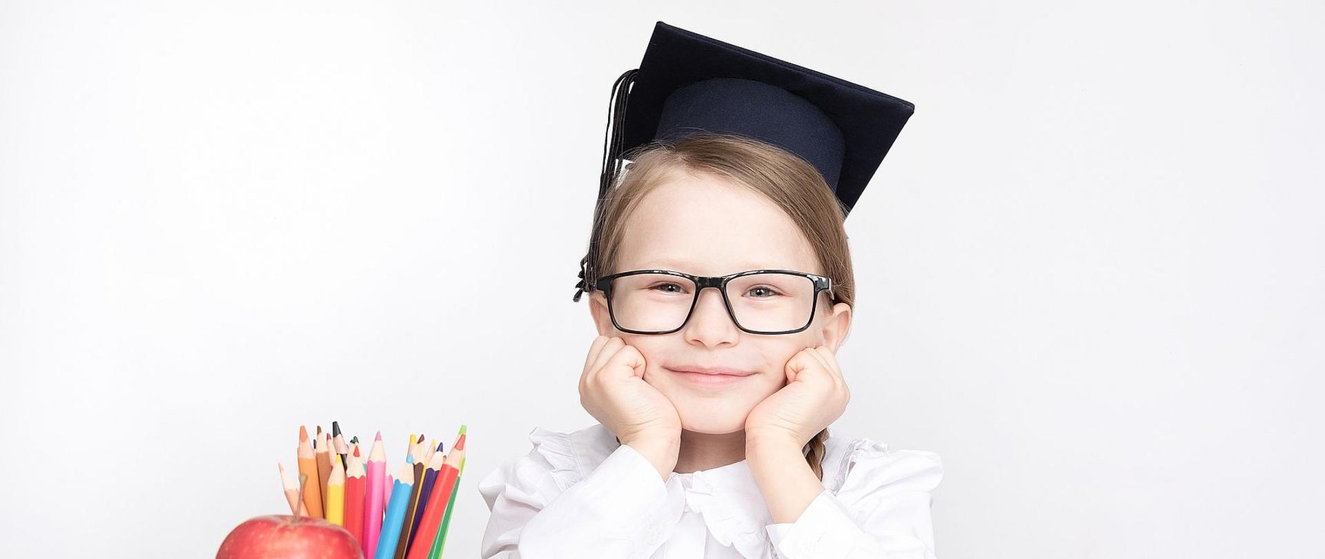 Zdjęcie przedstawia dziewczynkę w birecie siedzącą przy biurku szkolnym na którym stoją zeszyty, kubek z kredkami i jabłko. Dziecko ubrane jest w białą koszulę.