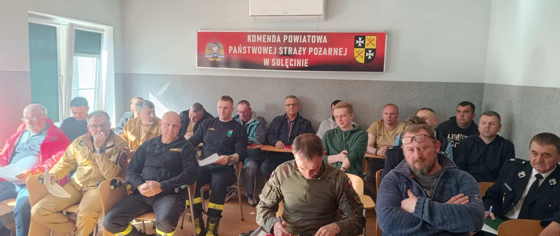 Strażacy OSP w sali na krzesłach na tle logo PSP Sulęcin