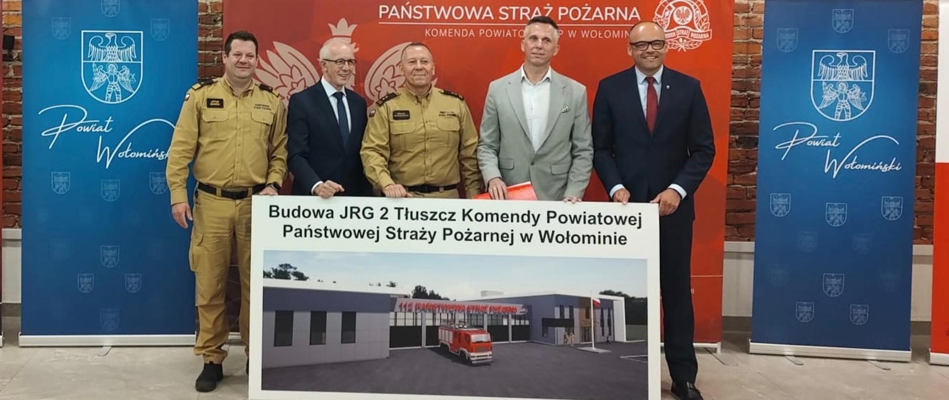 Na zdjęciu pięć osób stoi podczas uroczystości związanej z budową Jednostki Ratowniczo-Gaśniczej PSP w Tłuszczu. Dwie osoby ubrane są w mundury Państwowej Straży Pożarnej, pozostałe w stroje formalne. Uczestnicy prezentują planszę z wizualizacją przyszłej siedziby JRG 2 Tłuszcz Komendy Powiatowej PSP w Wołominie. W tle znajdują się banery Państwowej Straży Pożarnej, Powiatu Wołomińskiego oraz partnerów przedsięwzięcia. Uroczystość odbywa się we wnętrzu budynku z ceglaną ścianą.