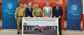 Na zdjęciu pięć osób stoi podczas uroczystości związanej z budową Jednostki Ratowniczo-Gaśniczej PSP w Tłuszczu. Dwie osoby ubrane są w mundury Państwowej Straży Pożarnej, pozostałe w stroje formalne. Uczestnicy prezentują planszę z wizualizacją przyszłej siedziby JRG 2 Tłuszcz Komendy Powiatowej PSP w Wołominie. W tle znajdują się banery Państwowej Straży Pożarnej, Powiatu Wołomińskiego oraz partnerów przedsięwzięcia. Uroczystość odbywa się we wnętrzu budynku z ceglaną ścianą.